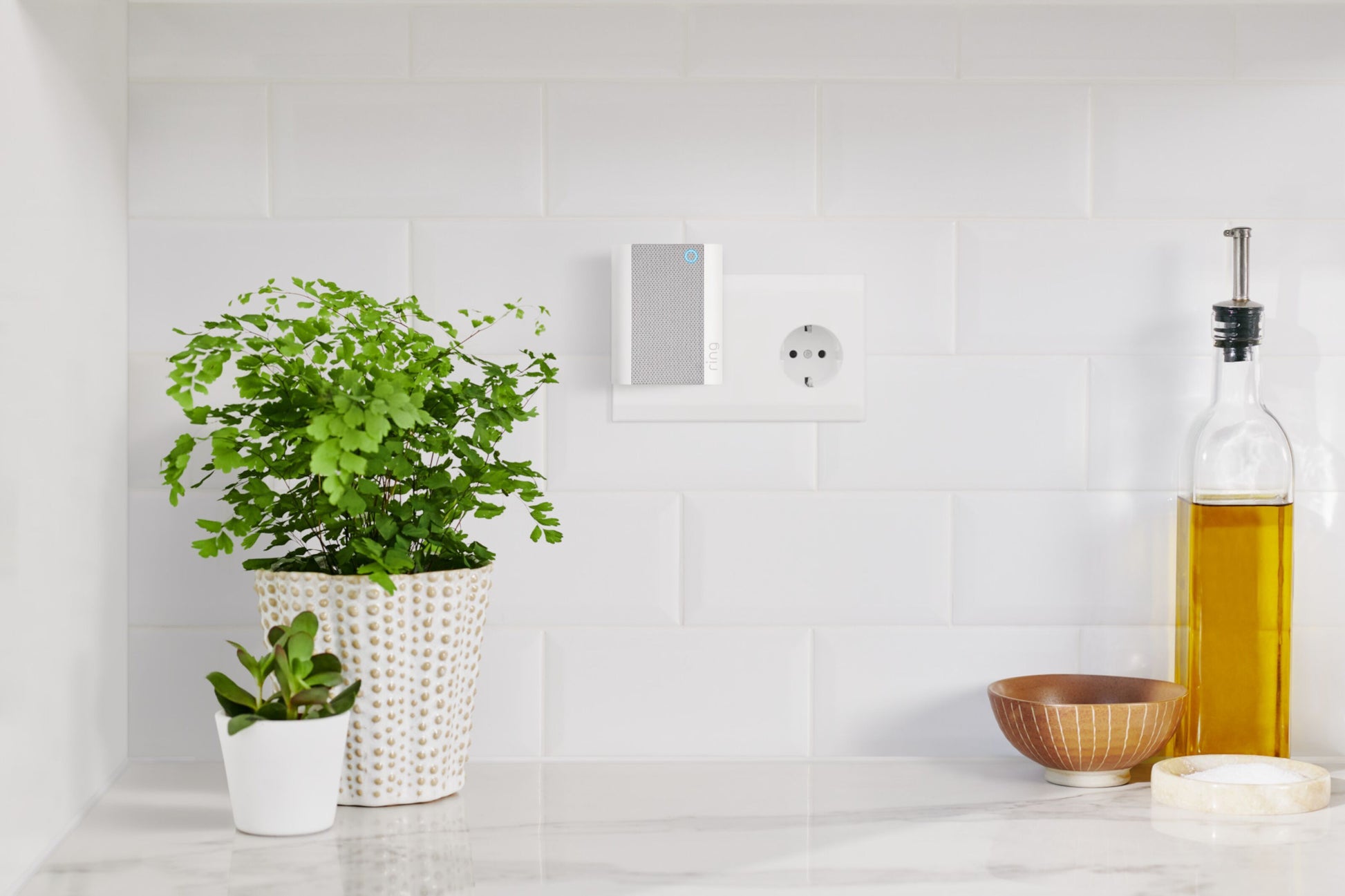 Ring Klokke Gen.2 hvid på køkkenbord med olieflaske, skål, urtepotteskjulere, stikkontakt og lille enhed.