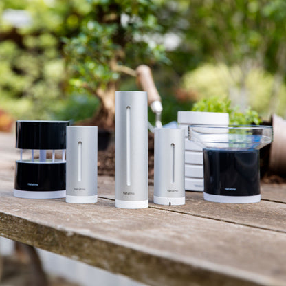 Netatmo Vejrstation Smart vejrstation i sølv på træbord udendørs ved planter og haveredskaber.