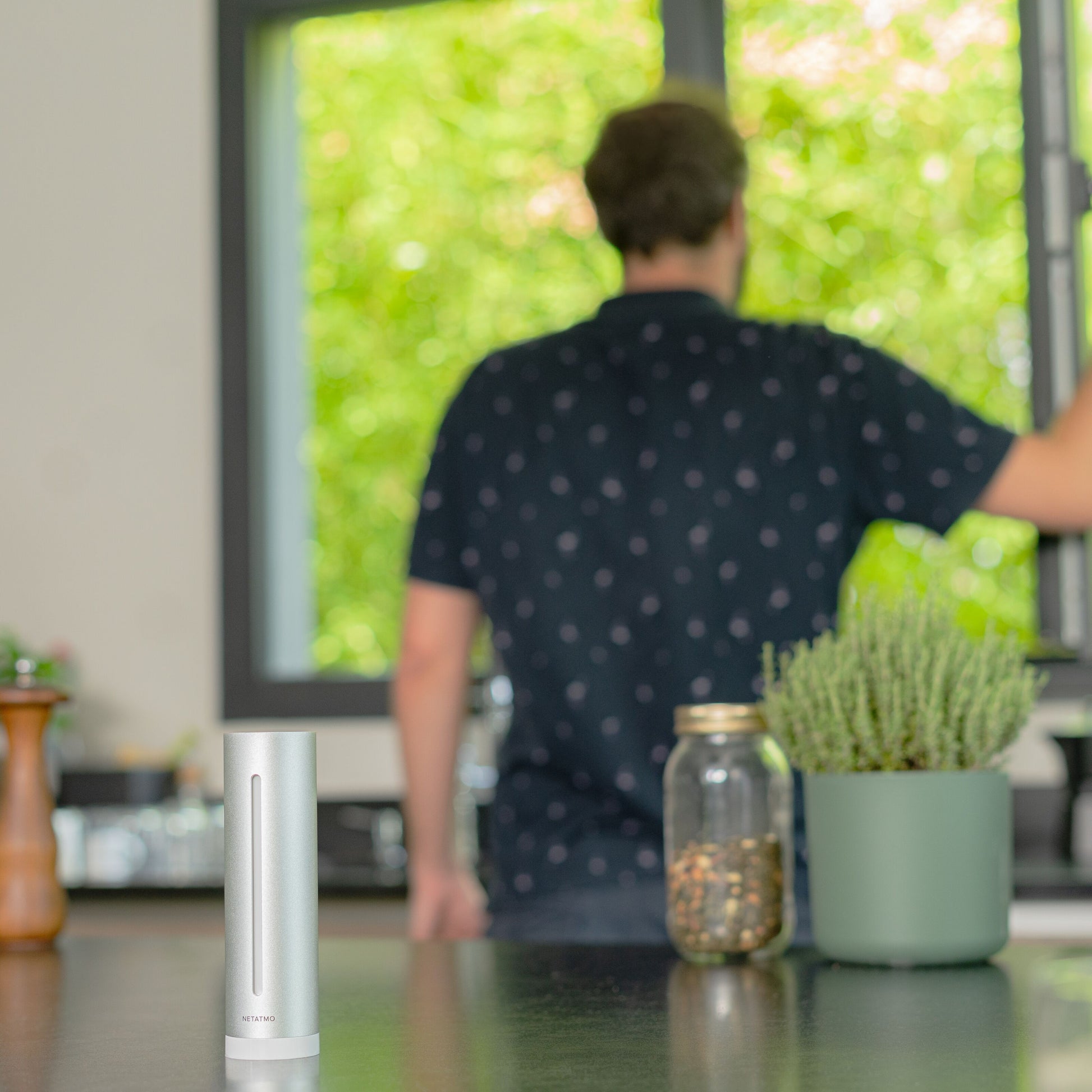 Netatmo Vejrstation Smart på køkkenbord ved krydderiglas og urtepotteskjuler, mand ved vindue i prikskjorte.