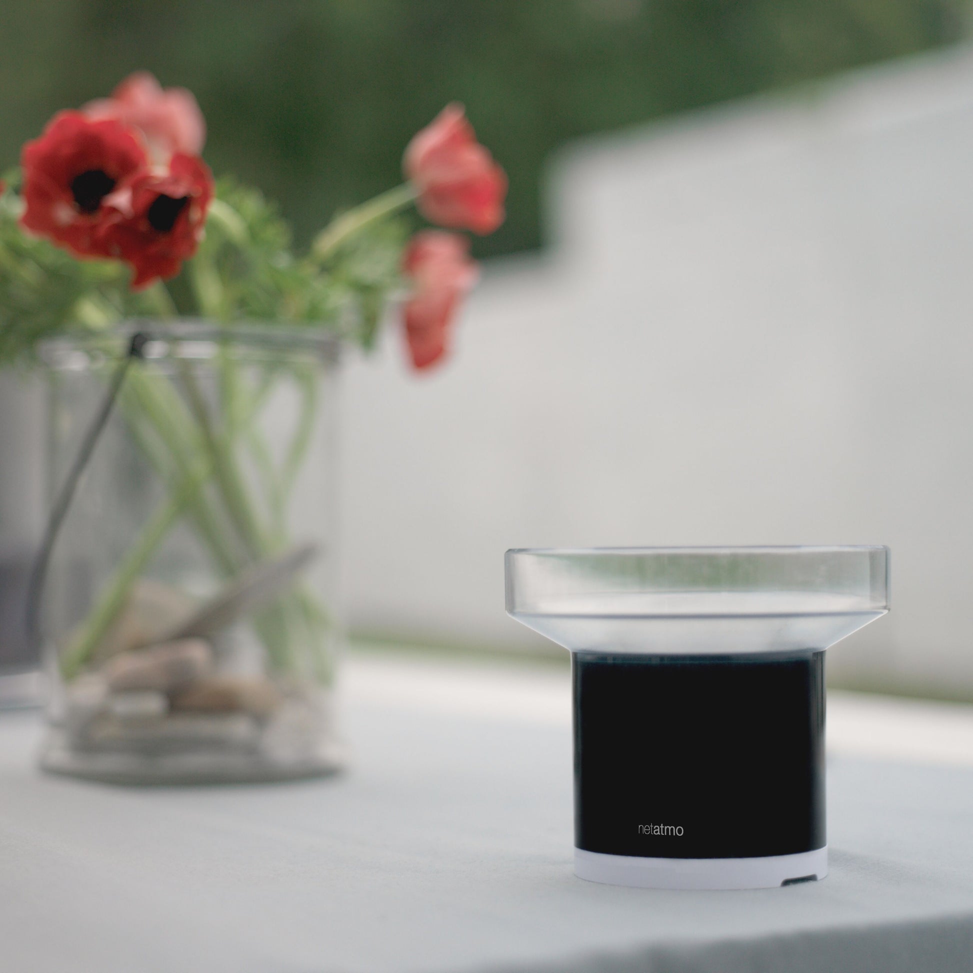 Netatmo Regnsensor sort/hvid til Netatmo vejrstation ved siden af glas med sten og røde blomster.