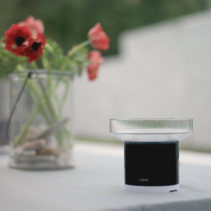 Netatmo Regnsensor sort/hvid til Netatmo vejrstation ved siden af glas med sten og røde blomster.