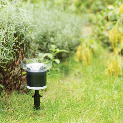 Netatmo Regnsensor sort/sølv til Netatmo vejrstation placeret i græs mellem grønne planter og gule blomster.