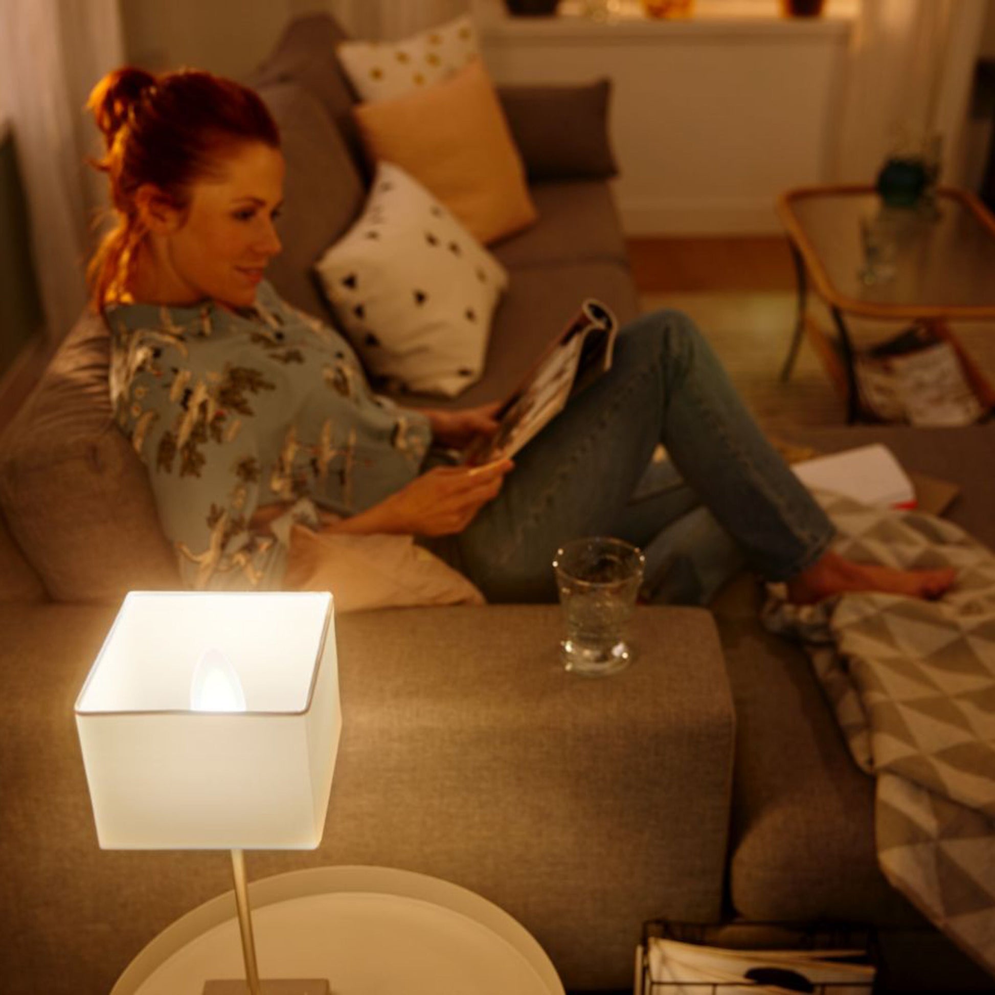 Philips Hue Ambiance Smart LED-pære E14 470 lm lyser stue med grå sofa, kvinde og glas vand på lille bord.