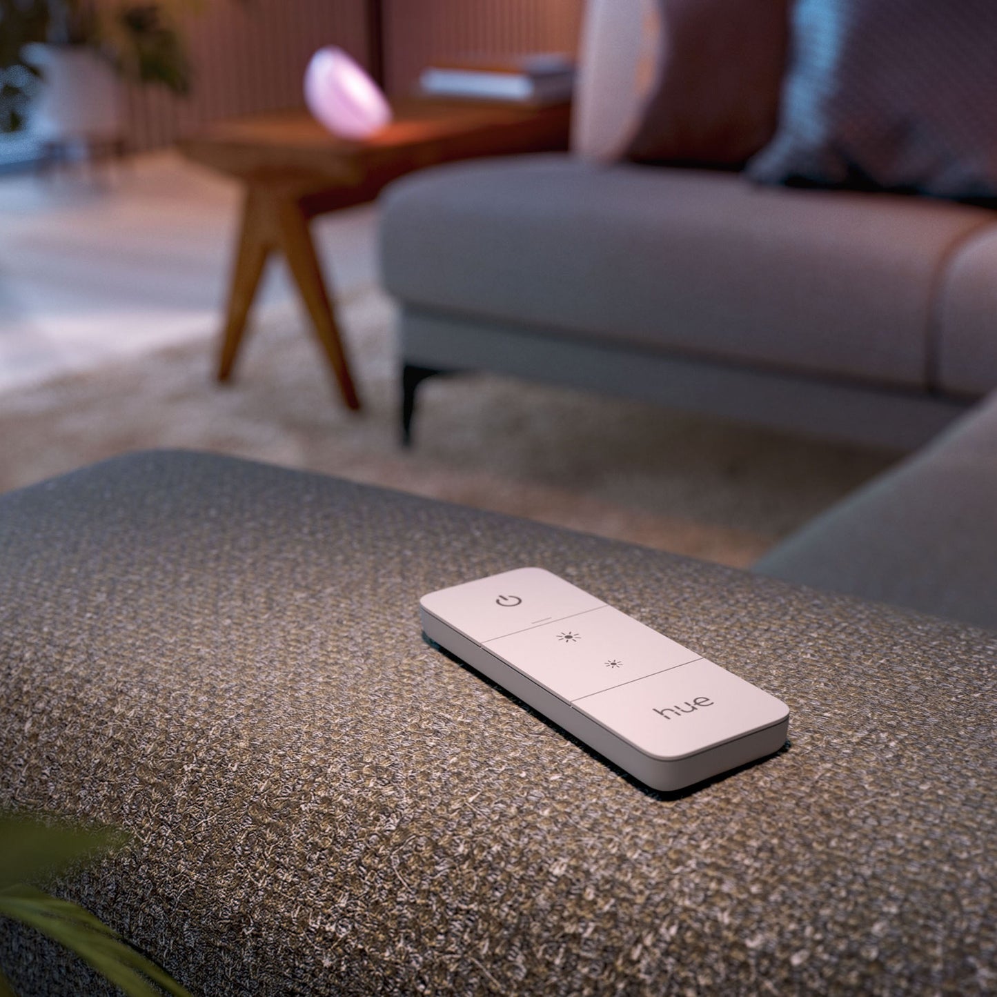 Philips Hue Dim Switch v2 fjernbetjening på grå puf foran sofa og træbord i stue med varm belysning.