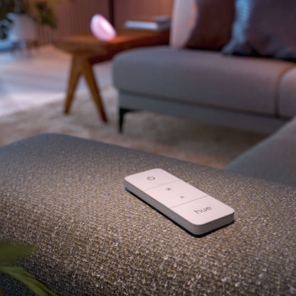 Philips Hue Dim Switch v2 fjernbetjening på grå puf foran sofa og træbord i stue med varm belysning.