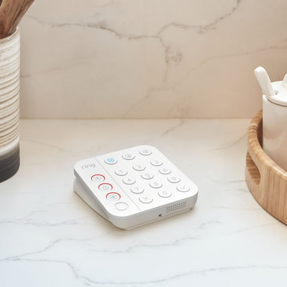 Ring Alarm 2. gen alarmsystem keypad på bordplade ved siden af keramisk beholder og fletkurv, hvid model.