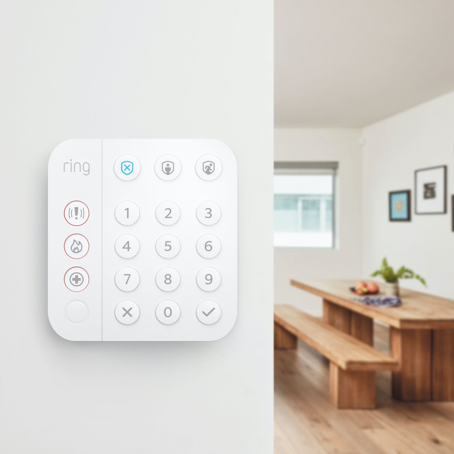 Ring Alarm tastatur monteret på hvid væg, trægulv synligt i spiseområde, bænke i baggrunden.
