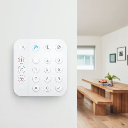 Ring Alarm tastatur monteret på hvid væg, trægulv synligt i spiseområde, bænke i baggrunden.