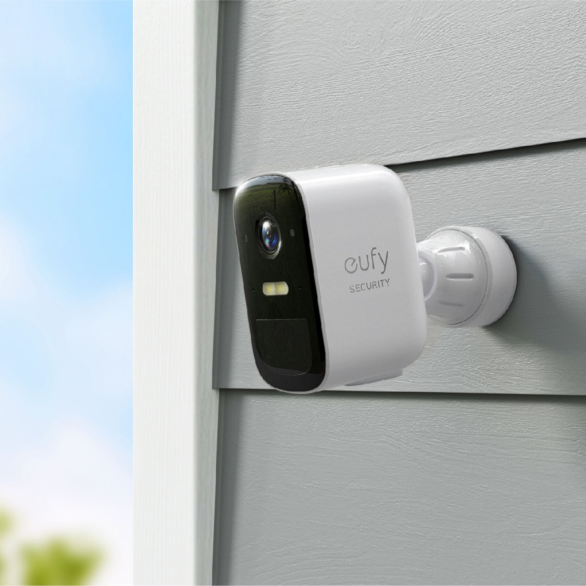 EufyCam 2C Ekstra kamera, hvid, monteret på grå facade og vinklet nedad, sløret blå himmel og træer bagved.