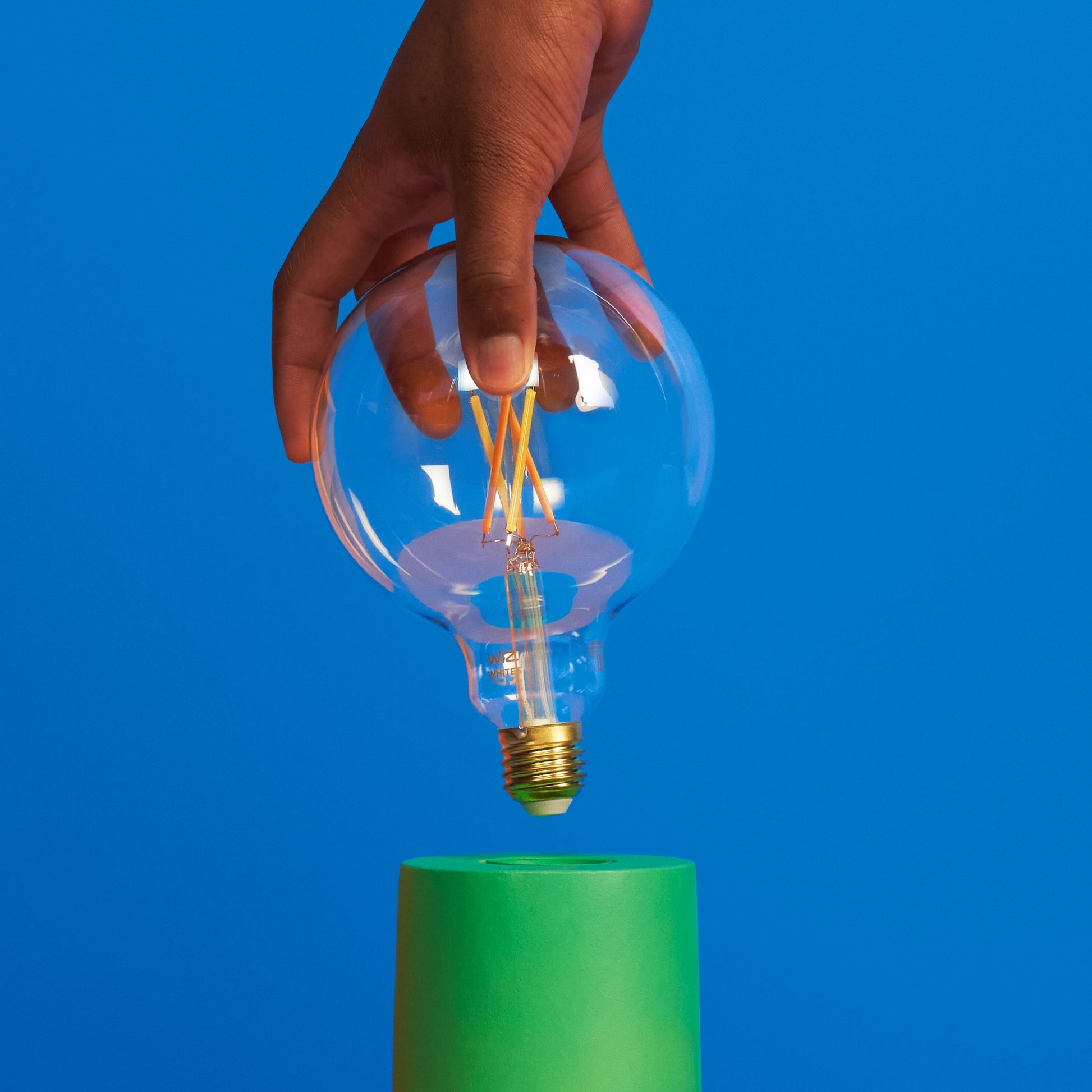 WiZ Klar Filament G95 Smart LED-pære E27 806 lm holdes over grøn cylinder, blå baggrund.