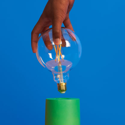 WiZ Klar Filament G95 Smart LED-pære E27 806 lm holdes over grøn cylinder, blå baggrund.