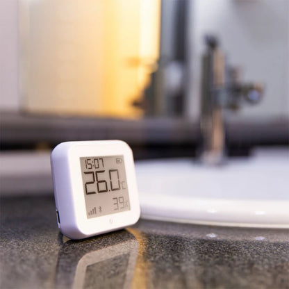 Shelly HT Gen 3 termometer og hygrometer hvid viser 26,0°C og 39 luftfugtighed på badeværelsesbord.