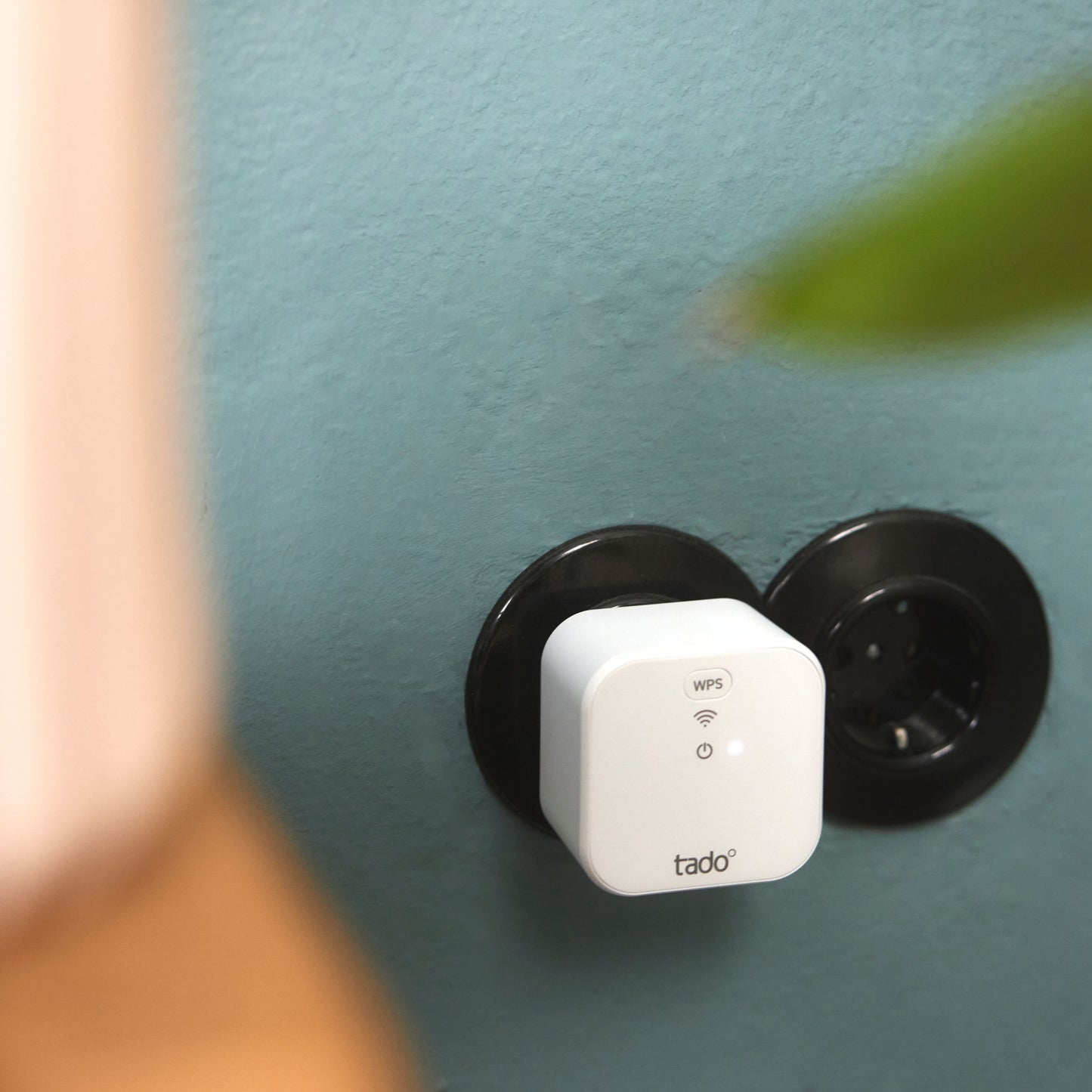 Tado Bro X grænserouter i sort stik på lyseblå væg, grøn bladkant og uklar lys genstand delvist synlig foran.