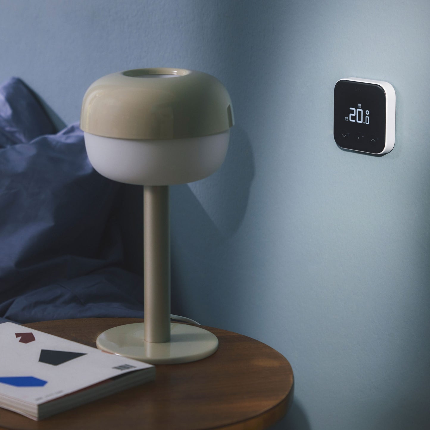 Tado Smart termostat X trådløs startpakke viser 20°C, monteret på blå væg over træbord med beige lampe og åben bog.