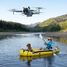 DJI Mini 3 drone optager 4K vertikale billeder af person og hund i gul gummibåd på flod med bjerge i baggrunden.