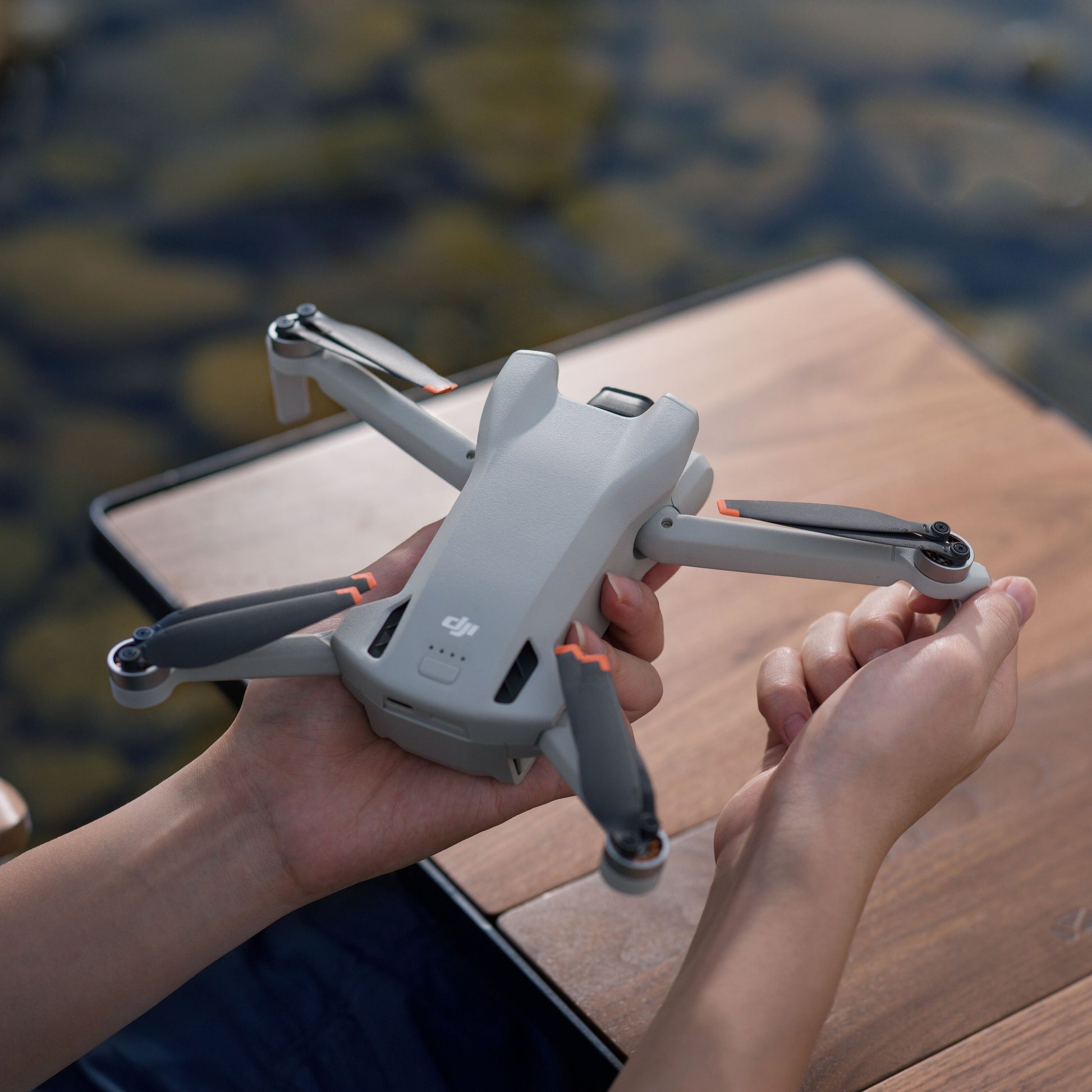 DJI Mini 3 grå drone med foldede arme holdes i hånden over træbord ved vand, sten ses under overfladen.