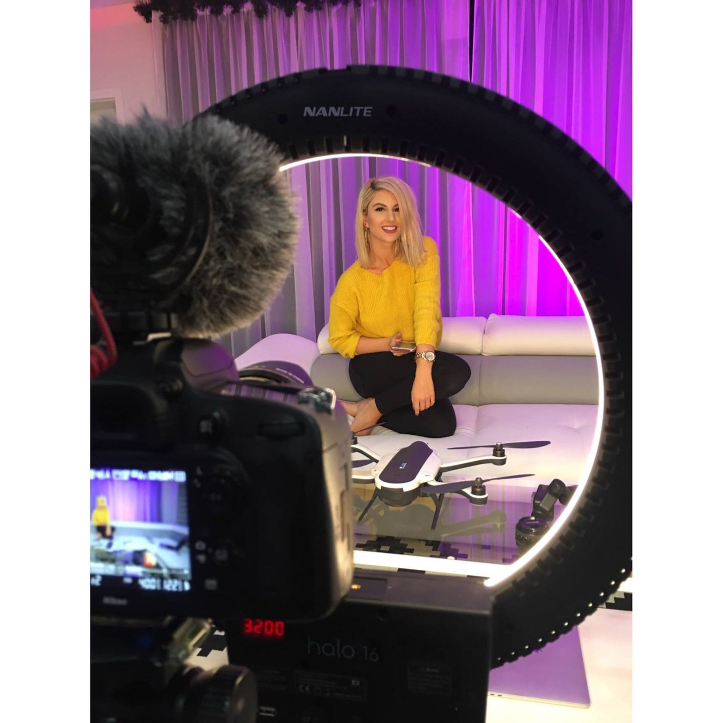 Nanlite Halo 16 LED-ringbelysning foran kvinde, kamera med mikrofon til livestream, drone på glasbord, lilla gardiner.