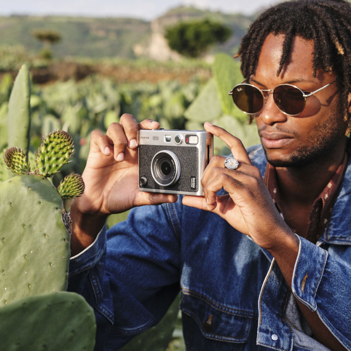 Fujifilm Instax Mini Evo Sort instantkamera bruges af person med solbriller og denimjakke blandt grønne kaktusser udendørs.