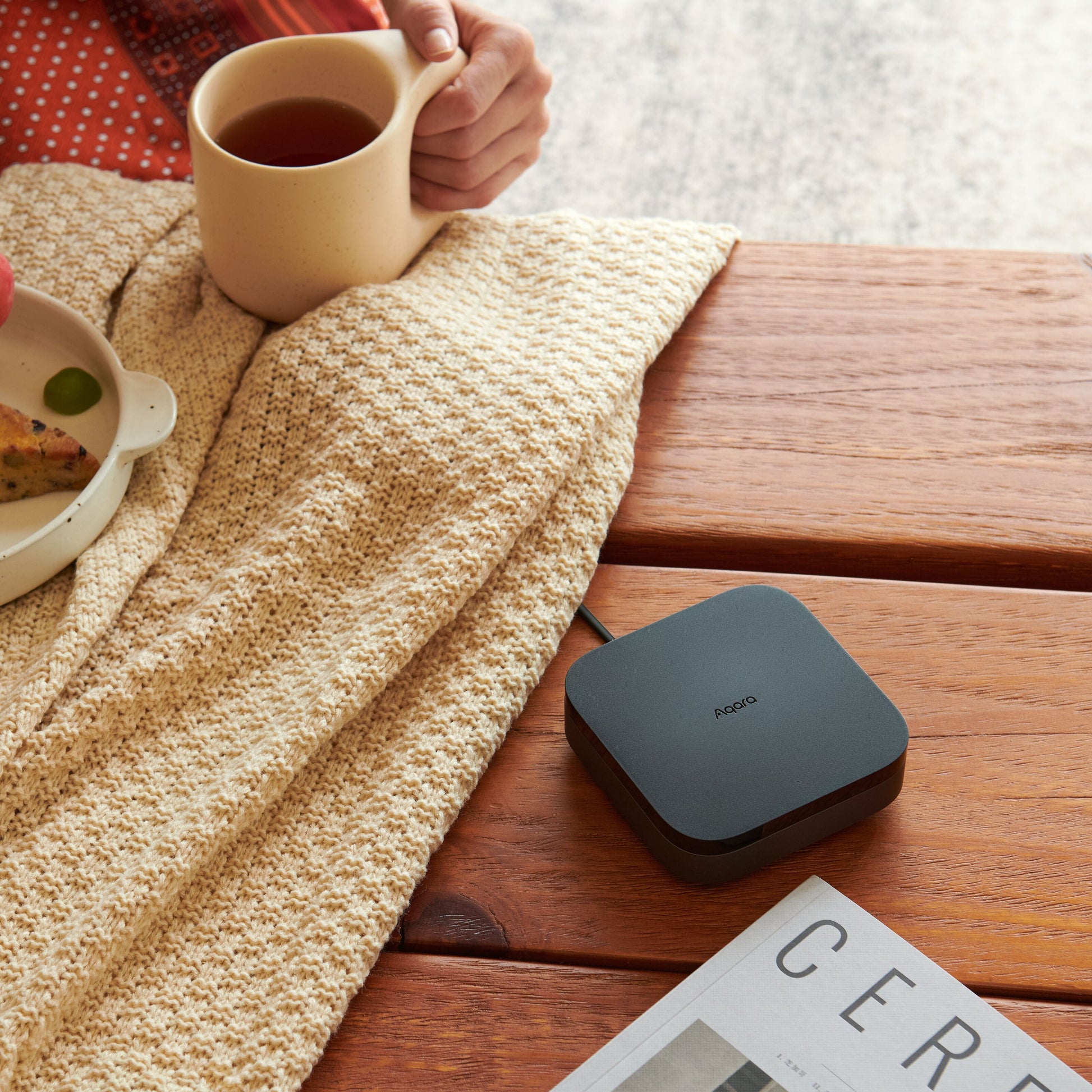 Aqara Hub M3 Smart Home Controller ved siden af sort Poco enhed, beige strikket tæppe, magasin delvist synlig på bord.