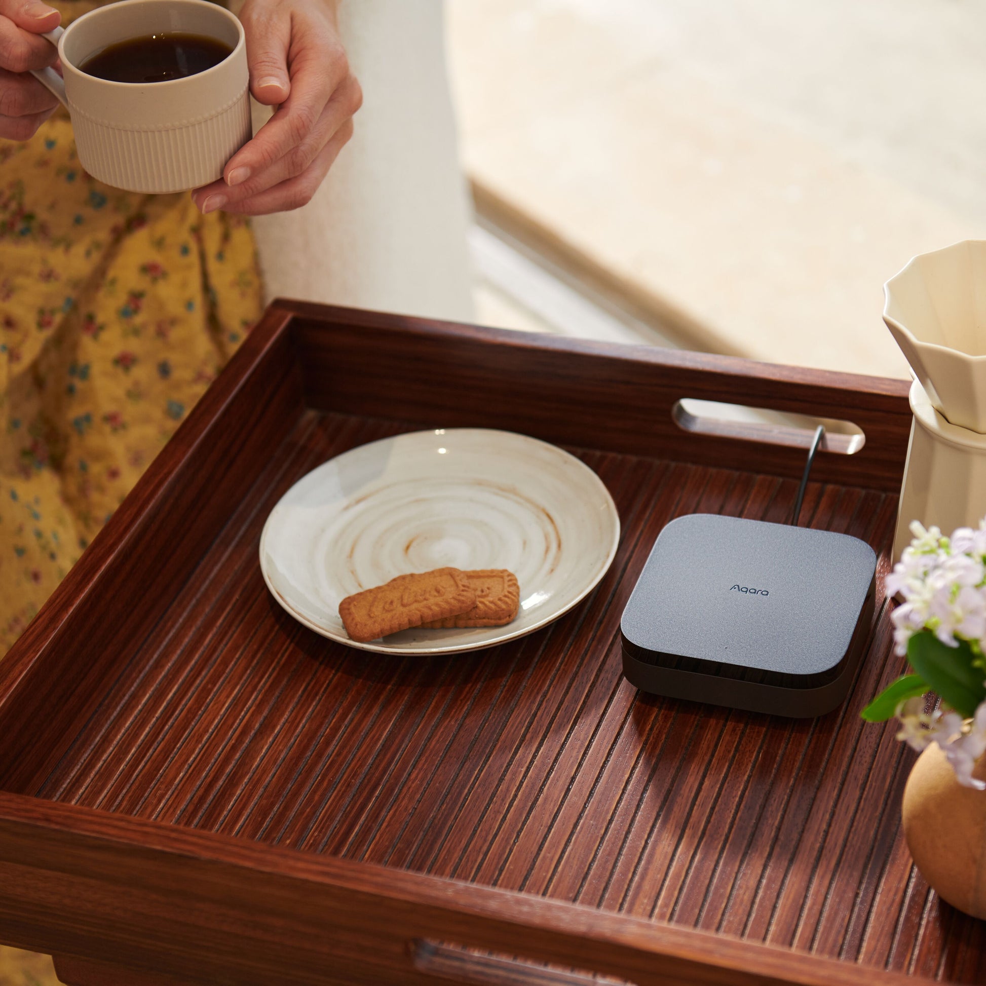 Aqara Hub M3 Smart Home Controller på træbakke ved siden af kop kaffe, to småkager og vase med blomster.
