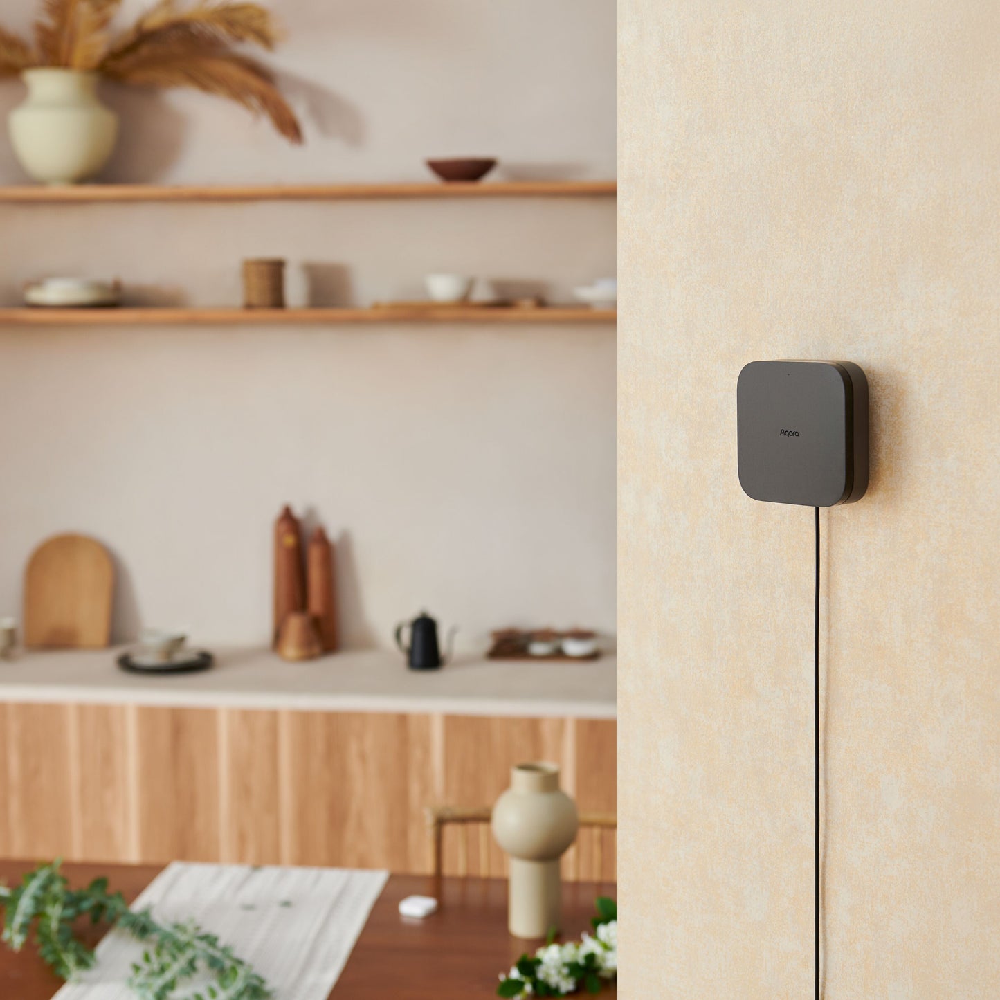 Aqara Hub M3 smart home controller i kvadratisk sort design med ledning monteret på lys væg, synlig del af rum og borde.