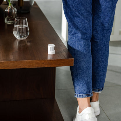 Aqara Bevægelses- og lyssensor AS5 på træbord med vandglas, person i blå jeans og hvide sneakers ved siden af.
