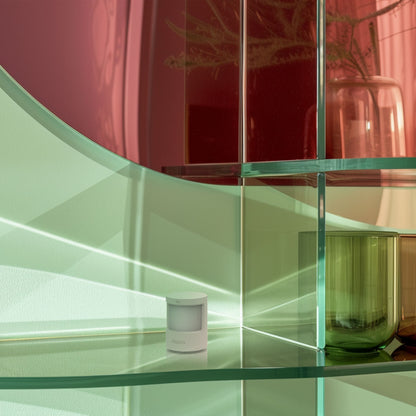 Aqara Bevægelses- og lyssensor AS5 på glashylde med glasvaser og plante synlig i baggrunden.