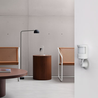 Aqara bevægelses- og lyssensor AS5 placeret på væg i rum med lysstole, træbord, lampe og tablet.