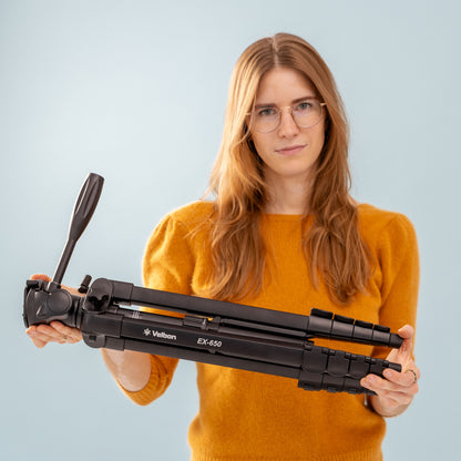 Velbon EX-650 stativ med mobilholder sammenfoldet vises, holdt af person i gul trøje, lys blå baggrund.