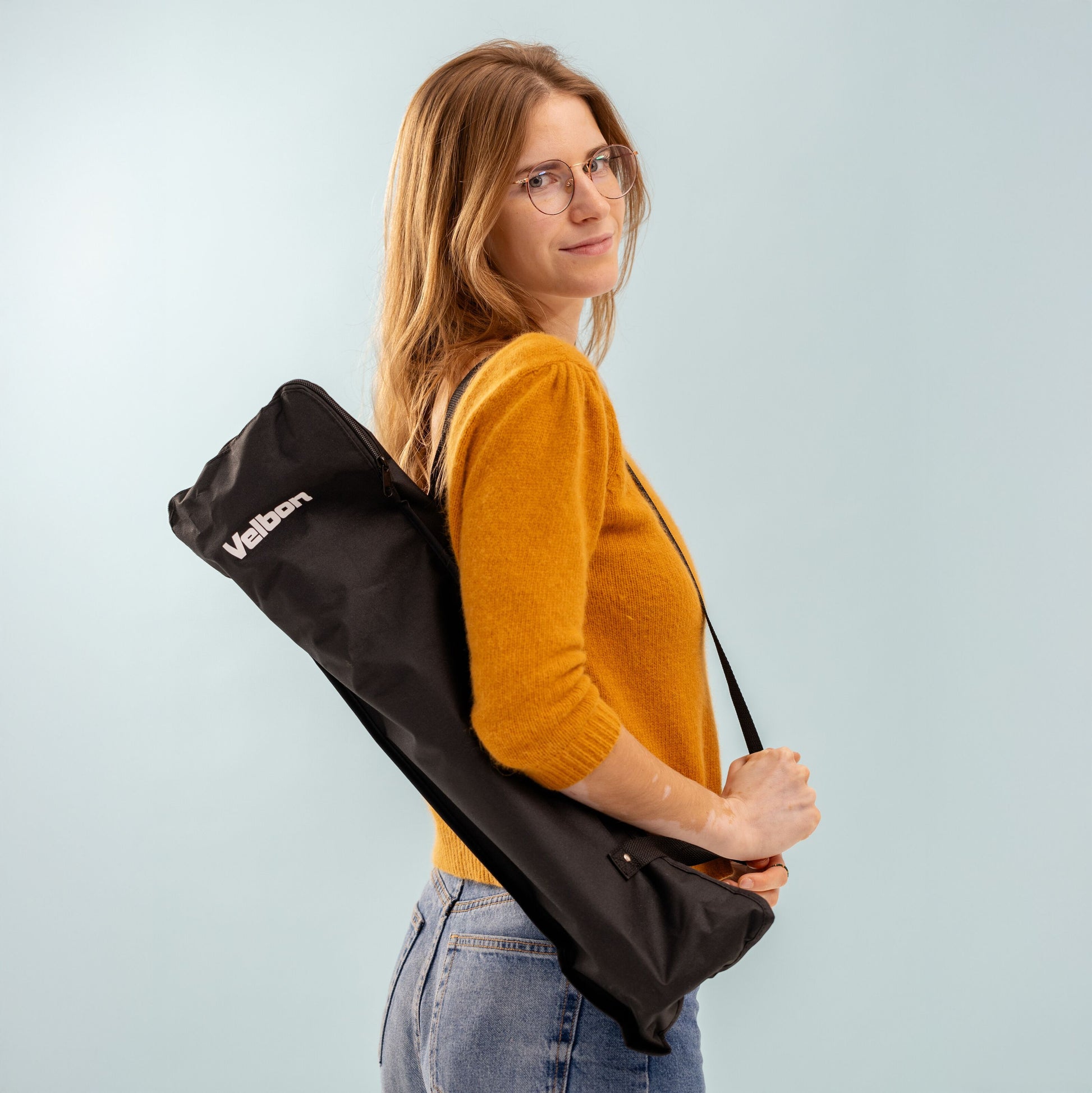 Velbon EX-650 med mobilholder bæretaske over skulder, kvinde i gul sweater og jeans, lys blå baggrund.