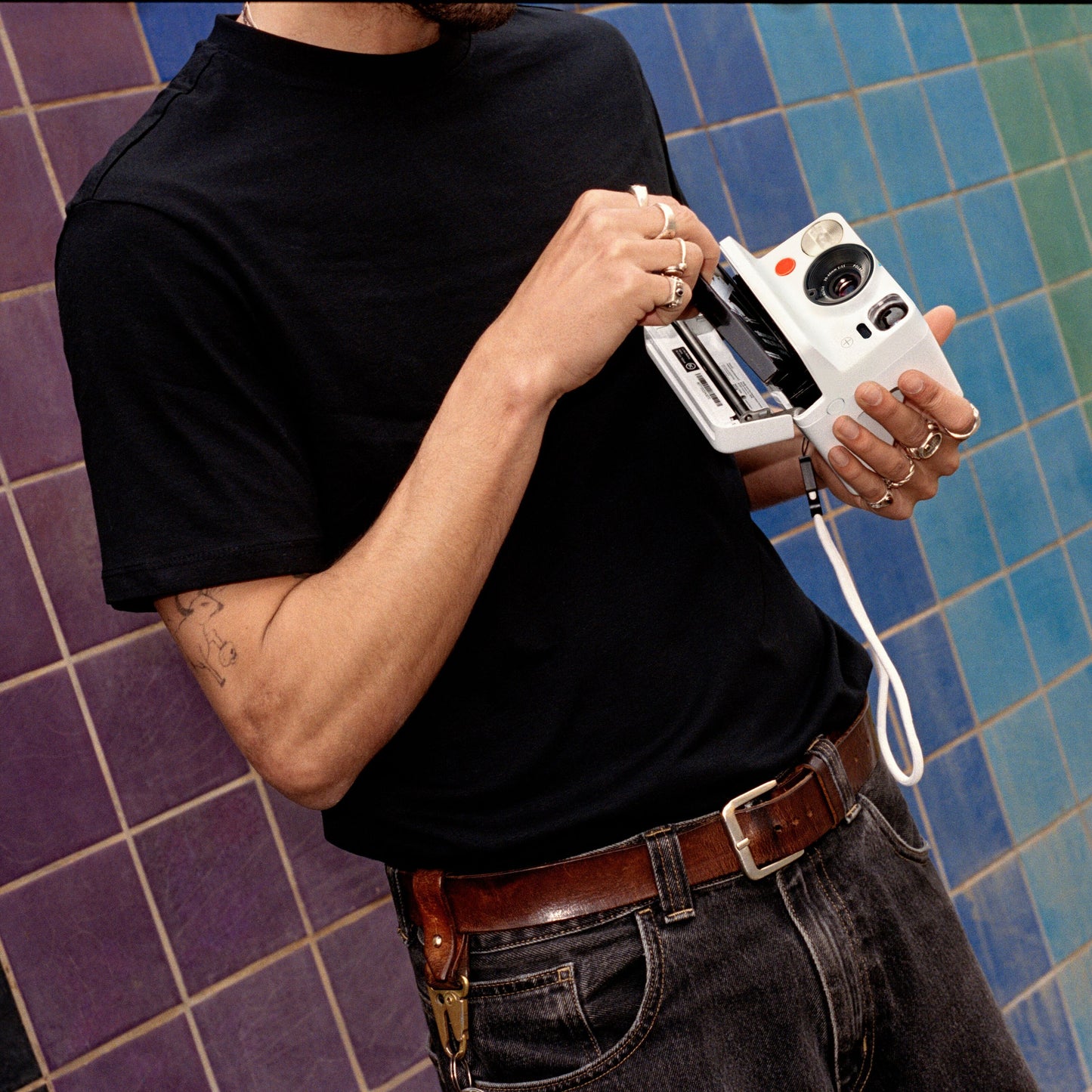 Polaroid Now Gen 3 sort kamera med film ses i brug af person i sort t-shirt og jeans foran blå-lilla flisevæg.