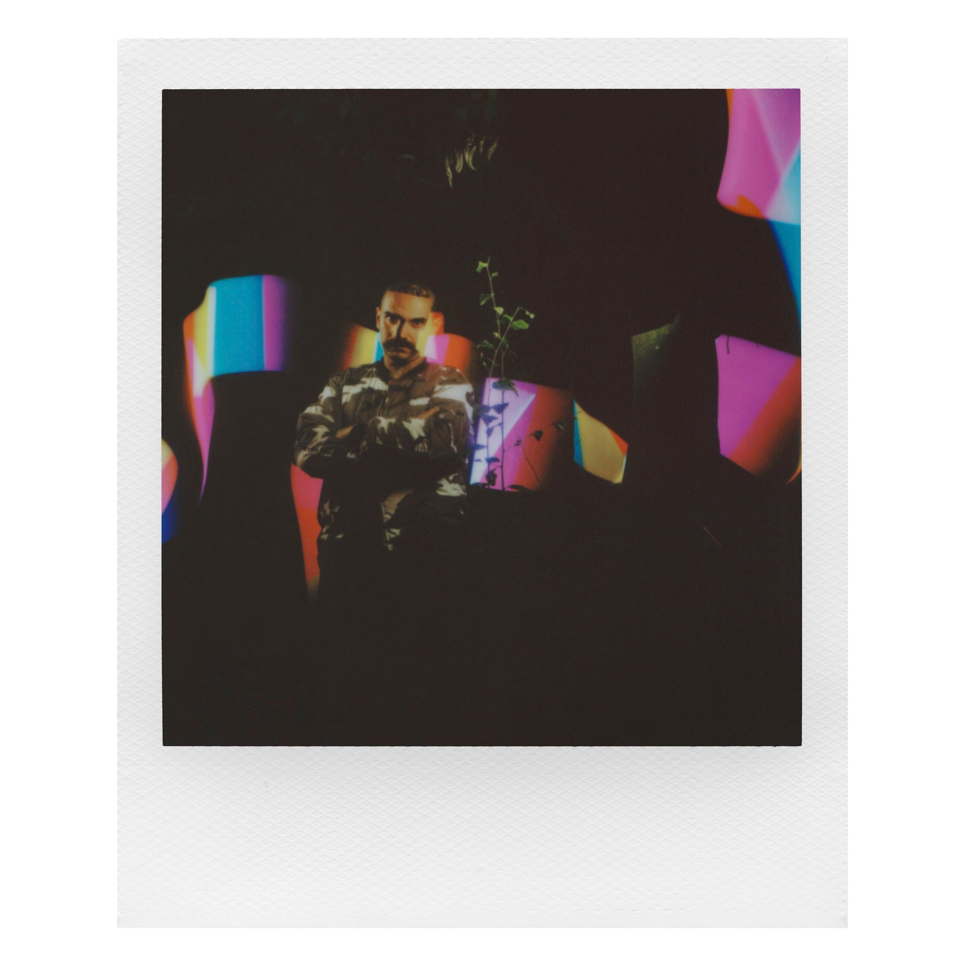 Polaroid Now Gen 3 Sort kamera med film, camouflage-skjorte, farvede former og plante i baggrunden.