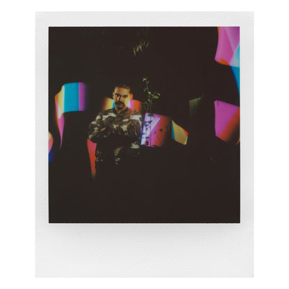 Polaroid Now Gen 3 Sort kamera med film, camouflage-skjorte, farvede former og plante i baggrunden.