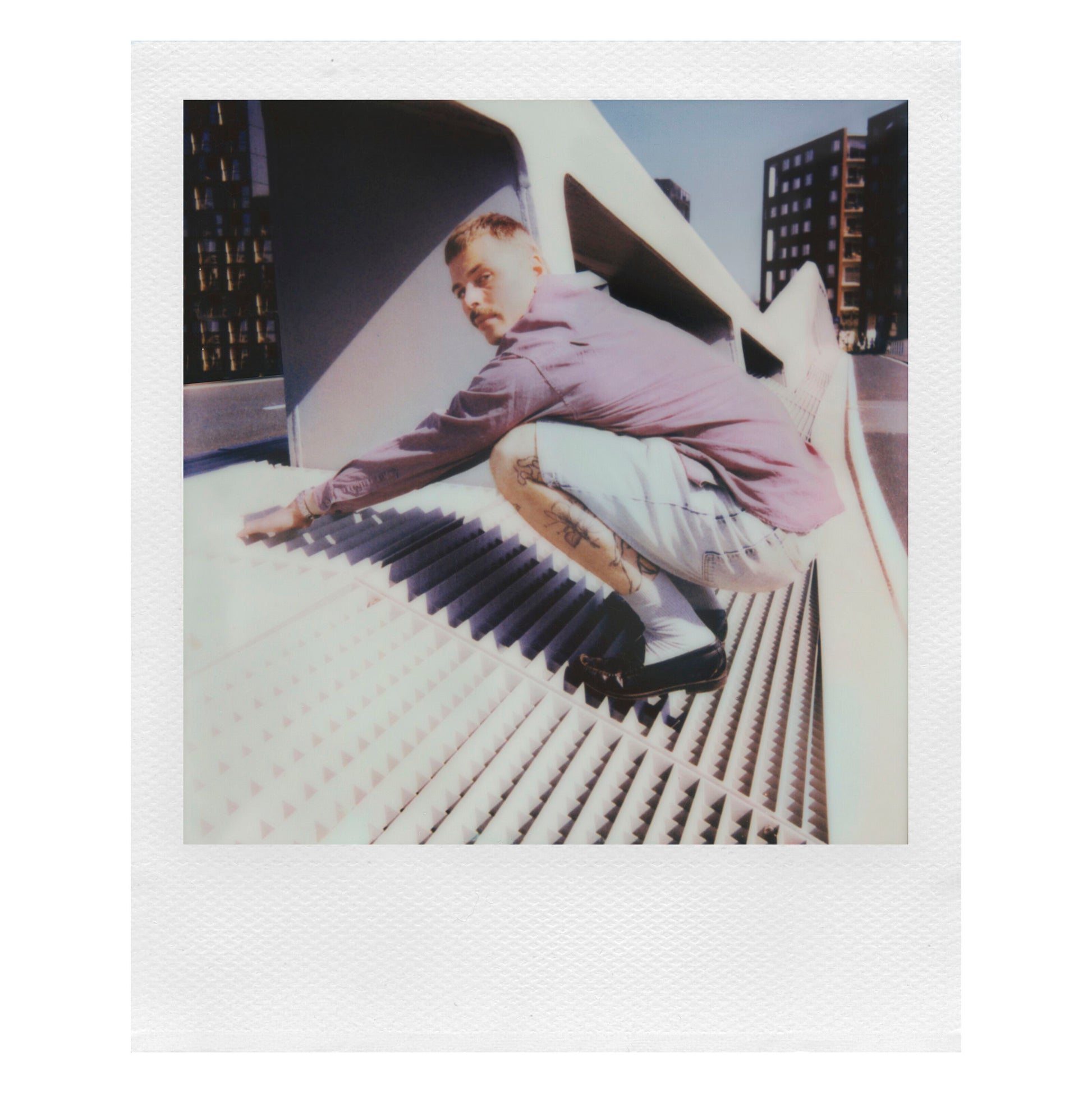 Polaroid Now Gen 3 Sort instant kamera med film holdes af mand i lyse lilla skjorte og denimshorts, synlige tatoveringer.