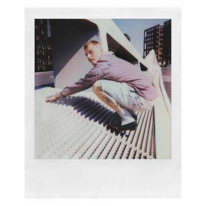 Polaroid Now Gen 3 Sort instant kamera med film holdes af mand i lyse lilla skjorte og denimshorts, synlige tatoveringer.