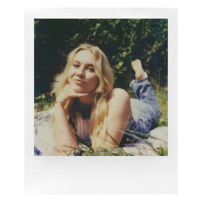Polaroid Now Gen 3 Sort med film ses i brug udendørs, kvinde med langt lyst hår på tæppe, omgivet af grønt.