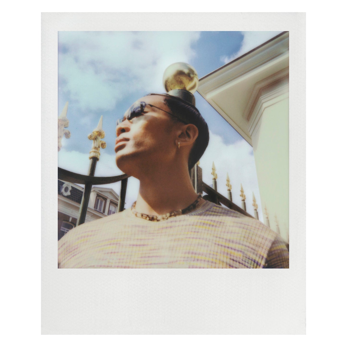 Polaroid Now Gen 3 Sort kamera med film, klassisk hvid ramme, ude ved gyldent hegn og bygning under skyet himmel.