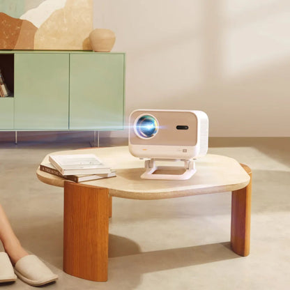 Yaber L2 Plus Full HD projektor på lys trækbord ved bøger og person, med JBL lyd, grøn skænk i baggrunden.