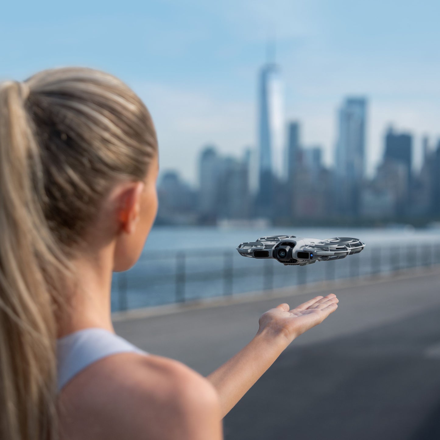 DJI Neo 2 drone i sort og sølv, flyvende over en udstrakt hånd, byskylinje og vand i baggrunden.