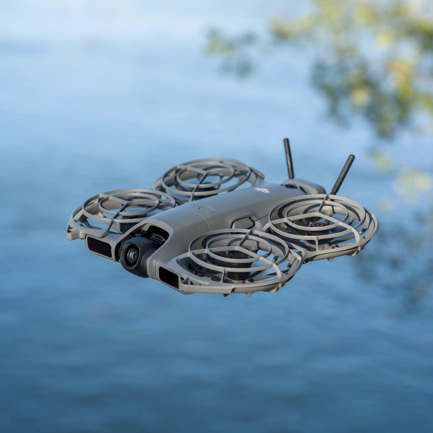 DJI Neo 2 grå ultralet drone med propelbeskyttere ses svævende over blåt vand med spejling af træer.