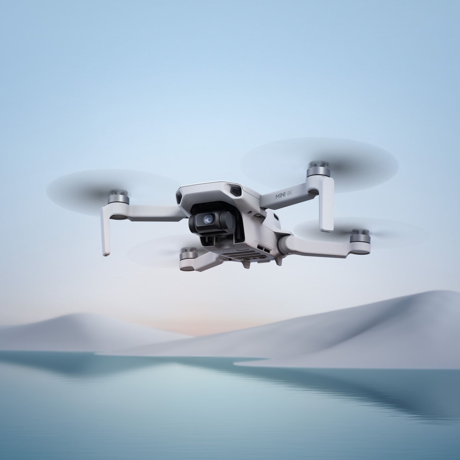 DJI Mini 4K Fly More Combo drone svæver over vand med sneklædte bakker i baggrunden, kompakt model med 4K-video.