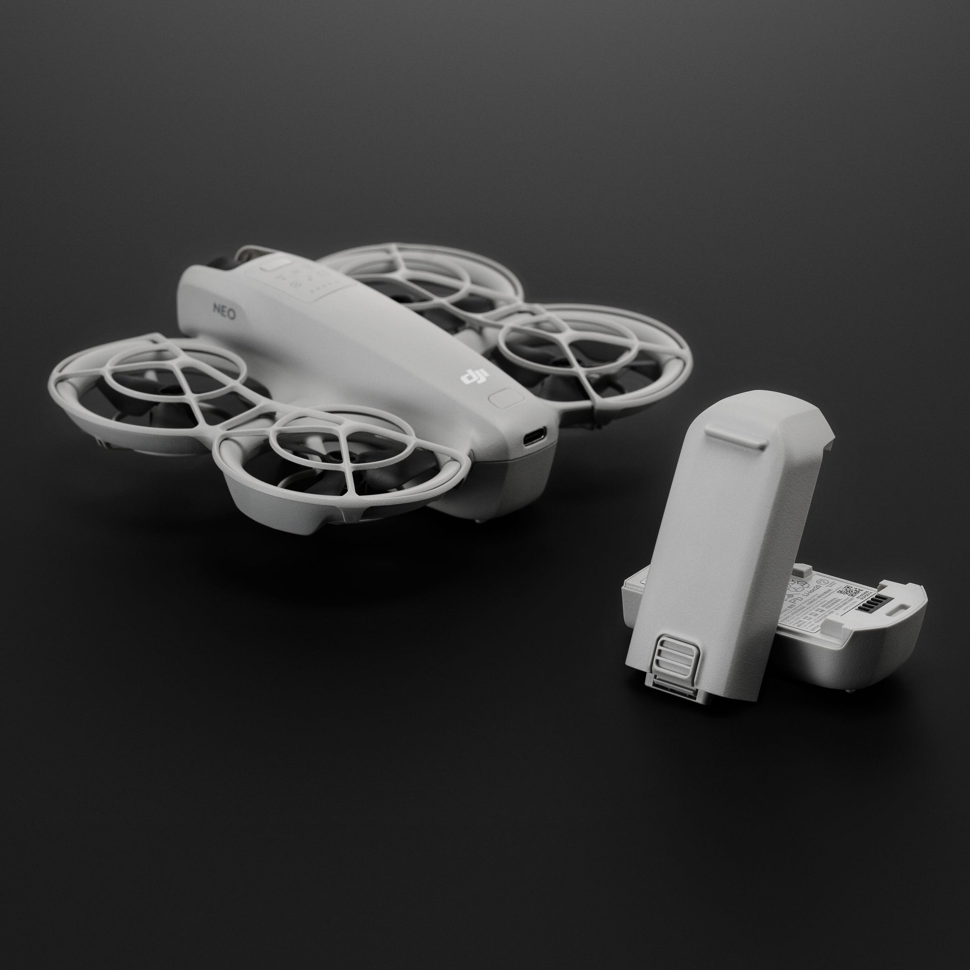 Grå DJI Neo drone med beskyttelsespropelbeskyttere og aftaget DJI Neo batteri på mørk overflade.