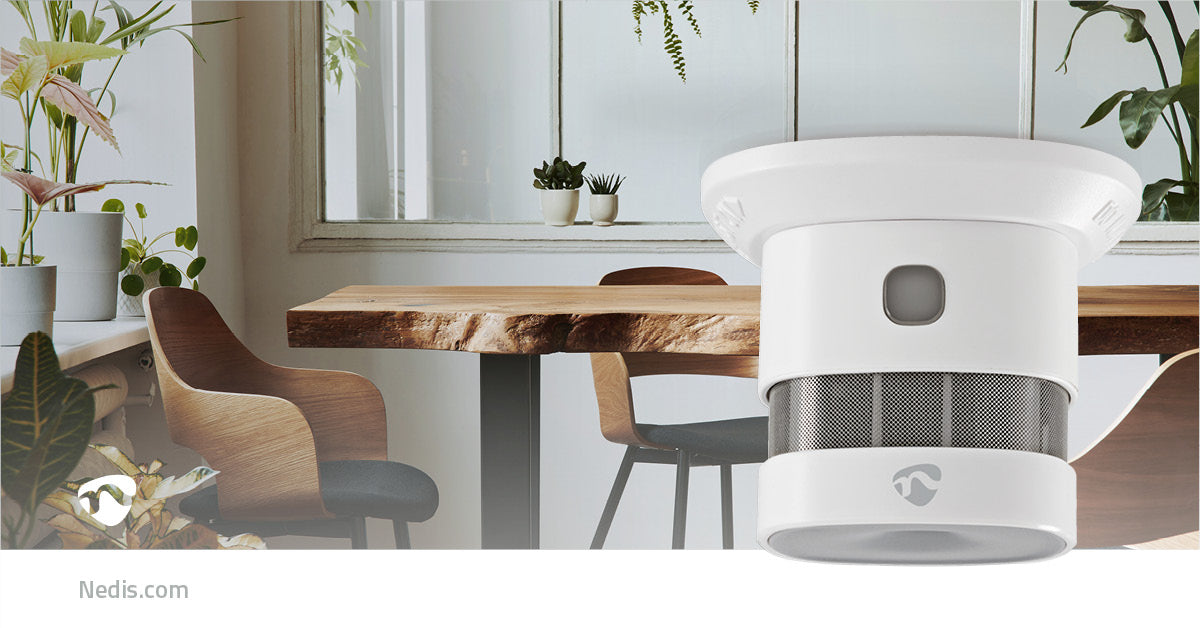 Nedis WiFi Brandalarm 5 år på bord i spiseområde med træmøbler og store vinduer, grønne planter i baggrunden.