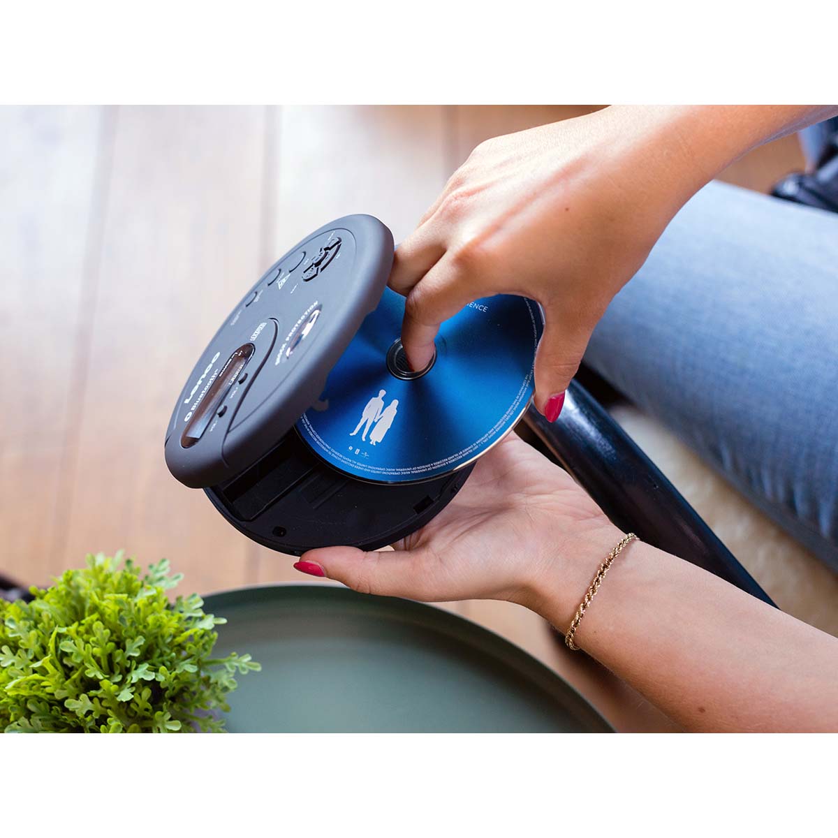 Lenco bærbar Bluetooth CD-MP3-afspiller sort, én hånd sætter blå CD i, grøn plante synlig i forgrund.