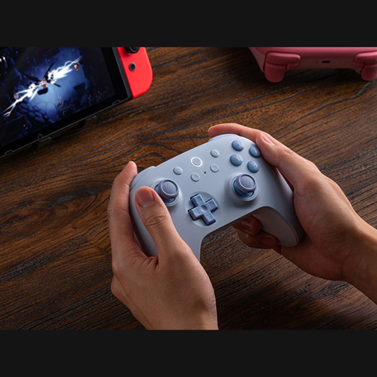Blå 8Bitdo Ultimate 2C Trådløs håndkontrol til Nintendo Switch holdes i hænder foran tablet med spil og rød enhed.
