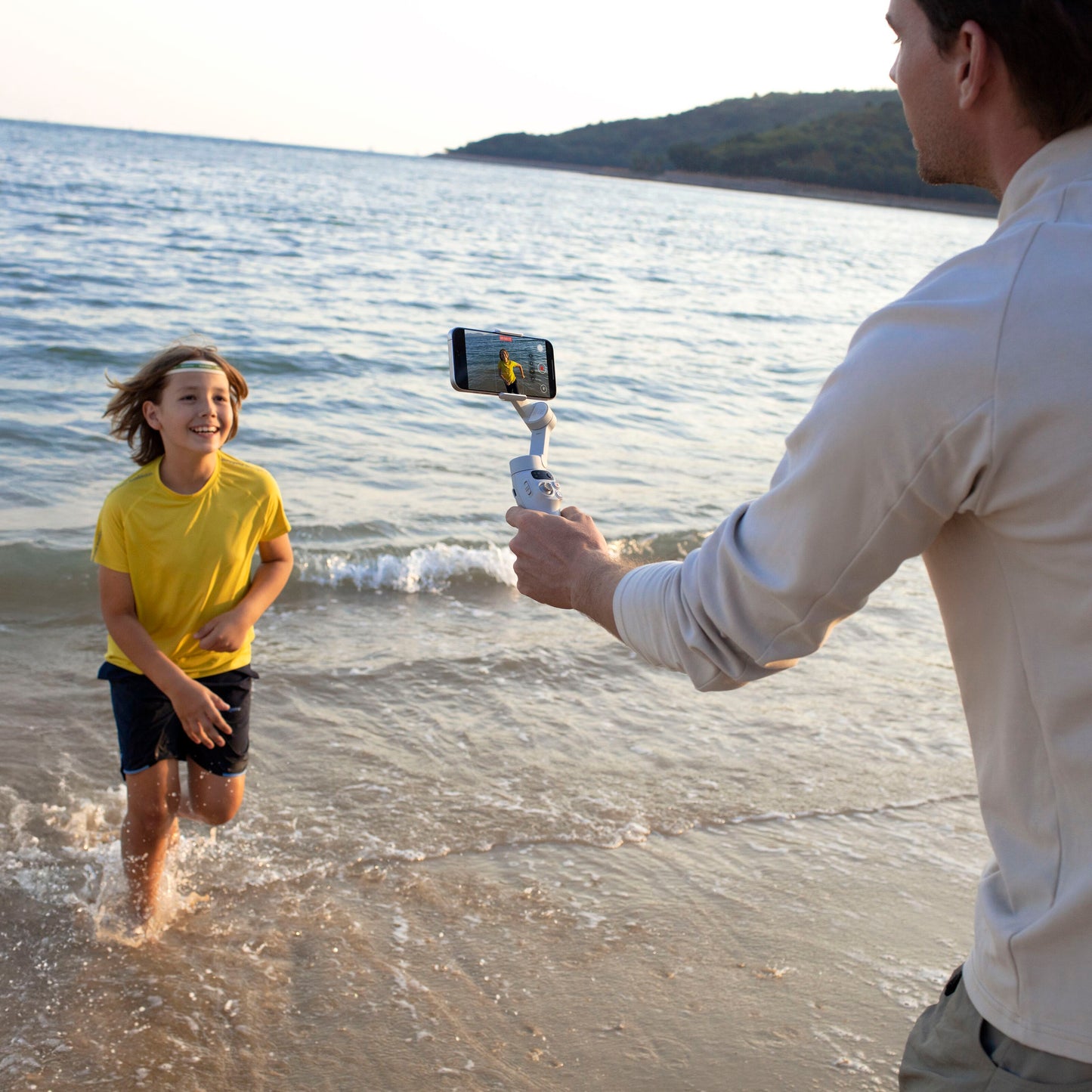 DJI Osmo Mobile 7 Gimbal stabiliserer smartphone under optagelse ved strand med løbende barn, vand og bakker i baggrund.