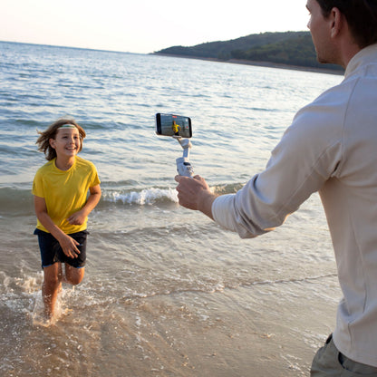 DJI Osmo Mobile 7 Gimbal stabiliserer smartphone under optagelse ved strand med løbende barn, vand og bakker i baggrund.