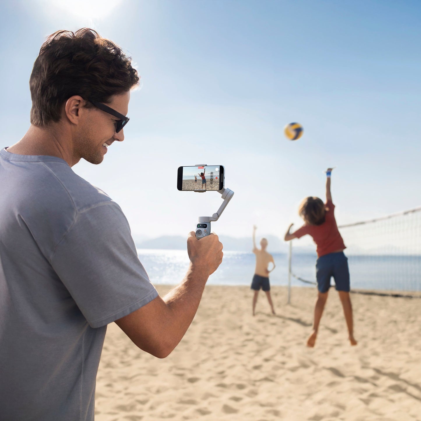 DJI Osmo Mobile 7 Gimbal sort i brug ved strand, mand filmer børn der spiller volleyball, blå himmel.