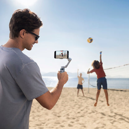 DJI Osmo Mobile 7 Gimbal sort i brug ved strand, mand filmer børn der spiller volleyball, blå himmel.