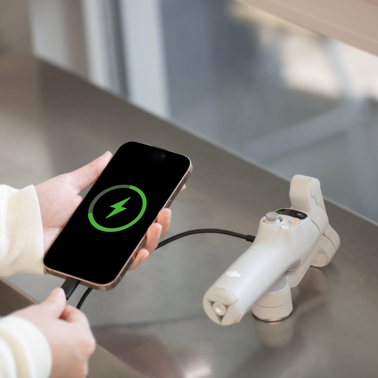 DJI Osmo Mobile 7 grå gimbal med smartphone forbundet via kabel, opladningssymbol synligt på telefonen.